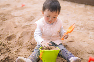 A toddler plays with sand, a green bowl, and an orange shovel outdoors.