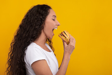 Obraz premium Smiling female model bites into a tasty burger with excitement. She is in a casual outfit against a vibrant yellow wall. Her joyful expression shows her love for this delicious fast food snack.