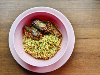 A simple budget-friendly meal of instant noodles topped with spicy canned fish, served in a pink bowl on a wooden table. Popular comfort food and an easy homemade dish in many Asian households.