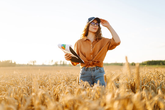 A female farmer wearing glasses stands in field with tablet in the sun's rays. Young female agronomist with tablet inspects harvest of golden wheat at sunset. Concept of gardening, agriculture. - Powered by Adobe