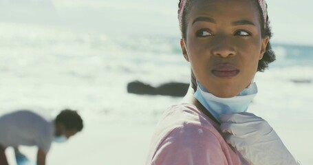 Adjusting woman pulling down surgical mask with gloved hand on sunny sandy shoreline, pink shirt