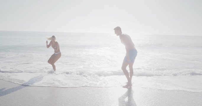 Jumping couple stepping back from incoming waves at shoreline, with bikini and blue swim trunks - Powered by Adobe