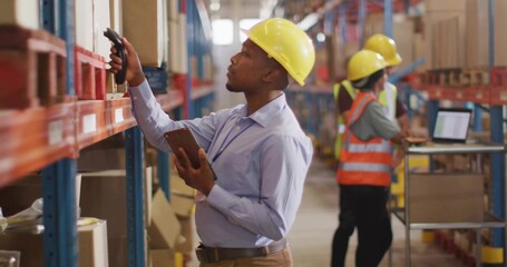 Inspecting warehouse supervisor wearing yellow hard hat scanning with barcode scanner and tablet