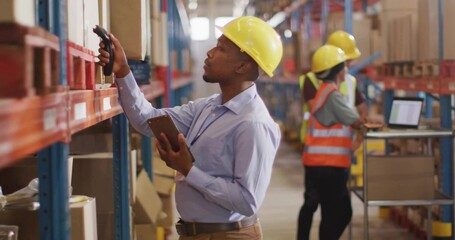 Reaching up warehouse picker wearing yellow helmet scanning box holding tablet and scanner in aisle
