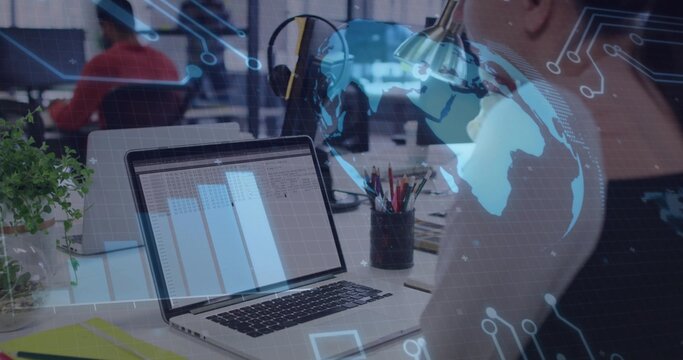 Reviewing laptop spreadsheet, woman wearing light top working at open office with pen cup and plant