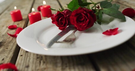 Displaying three red roses resting on plate with cutlery on weathered wooden table, 4 lit candles