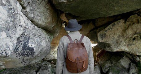 Standing hiker peering into crevice beneath overhang, wearing light jacket dark hat brown backpack