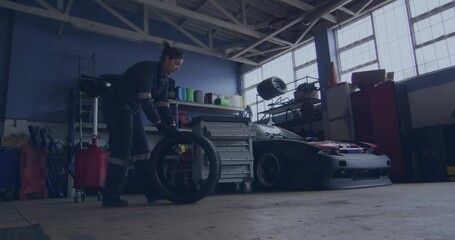 Bending female mechanic in dark coveralls handling spare tire by sports car at workshop, copy space