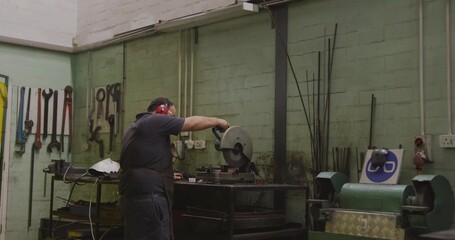 Operating metalworker cutting steel at small workshop with cut-off saw, red earmuffs, dark apron