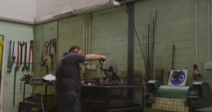 Operating metalworker cutting steel at small workshop with cut-off saw, red earmuffs, dark apron