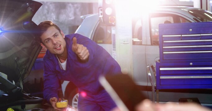 Leaning mechanic in blue coveralls checking open hood in garage, holding yellow tool near phone