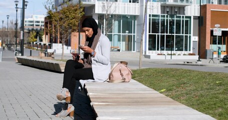 Sitting woman hijab cardigan checking phone holding takeaway cup on bench, pink bag, copy space