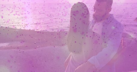 Hugging couple standing at waterline, woman in light dress, man in white shirt, pink speckle filter