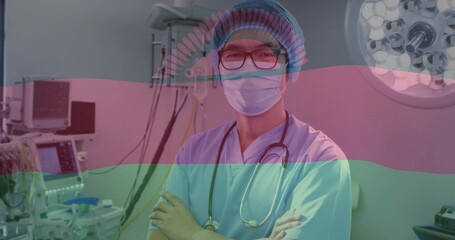 Standing clinician arms crossed wearing scrubs bouffant mask glasses in OR stethoscope and monitor