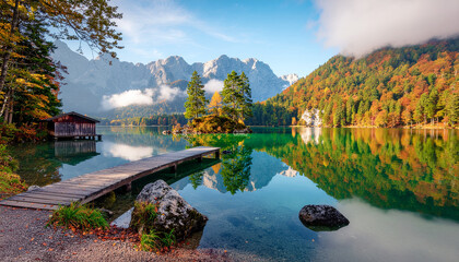 Serene view of Eibsee lake with turquoise water reflecting autumn forests and the Zugspitze massif. Features a small island, boathouse, and wooden pier.