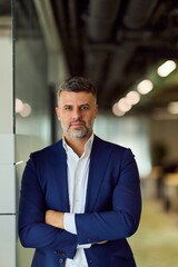 Obraz premium Confident Businessman In Navy Suit Stands Beside Glass Wall In Modern Office Corridor
