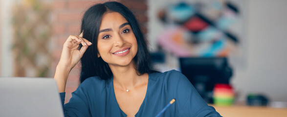 A young woman is sitting at a desk in a bright office. She is smiling and looking directly at the camera while holding a pen and appearing engaged in her work.