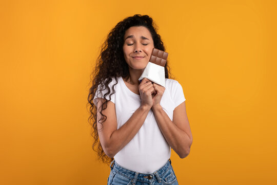 A joyful Latin lady is hugging a chocolate bar while closing her eyes in delight. She poses against a vibrant yellow-orange background, radiating hunger and love for cocoa.