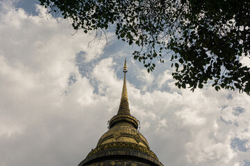 golden spire of a major Northern Thai (Lanna) temple stupa, likely the principal chedi of Wat Phra That Lampang Luang, framed dramatically by dark foliage and set against a vast, cloudy sky, symbolizi