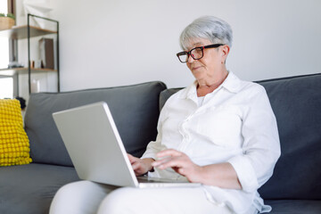 Mature woman with short gray hair and glasses works on laptop while sitting on sofa in sunny room. Beautiful woman sits relaxed on sofa with laptop in cozy room. Concept of happy aging, relaxation.