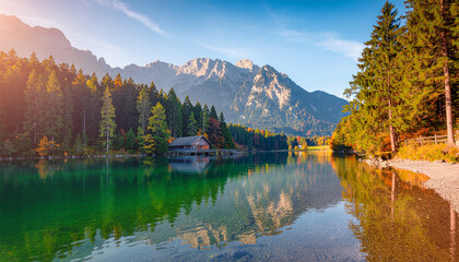 Clear alpine lake reflecting a wooden boathouse, vibrant autumn forest, and rugged mountain peaks under a bright sun. Tranquil nature scene.