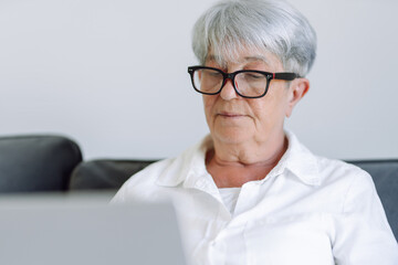 Mature woman with short gray hair and glasses works on laptop while sitting on sofa in sunny room. Beautiful woman sits relaxed on sofa with laptop in cozy room. Concept of happy aging, relaxation.