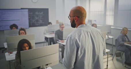 Professional Male Instructor Stands in Front of Adult Students in Computer Lab, Explaining Coding Principles. Concept Continuous Education, Digital Skills, and Modern Learning Environment. Back View.