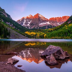 A scenic mountainous landscape mirrors in calm water with trees and rocks in the foreground and a brilliant sky above