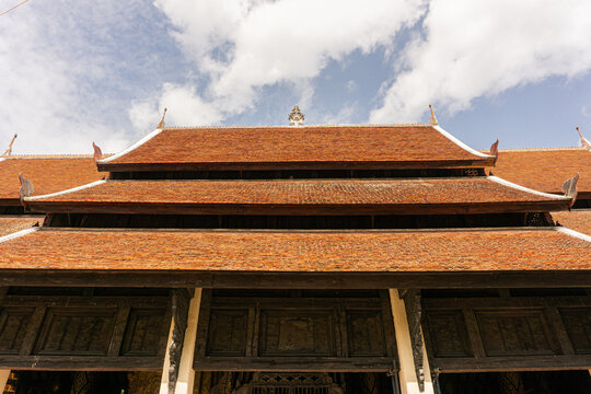 traditional Lanna style temple or hall. The dark wooden structure beneath supports the warm, reddish-brown layers of tiles, leading up to the pointed chofa (sky tassel) at the ridge. The composition h