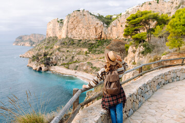 View from behind. Female hiker wearing hat and carrying backpack stands on edge of cliff with stunning sea view. Young woman feels free, enjoys nature on sunny day. Concept of hiking, freedom. 