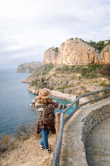 View from behind. Female hiker wearing hat and carrying backpack stands on edge of cliff with stunning sea view. Young woman feels free, enjoys nature on sunny day. Concept of hiking, freedom. 