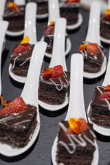 Bite sized dark chocolate brownies with glossy ganache, fresh strawberry, on white tasting spoons on a slate board