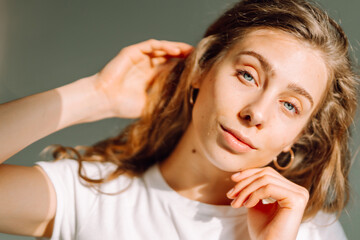 Naklejka premium A close-up of a young woman with acne on her face looks into a mirror in a sunny room. The beautiful woman enjoys her natural beauty reflected in the room. Concept of grooming and beauty.