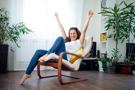 Happy woman relaxing with arms raised in a cozy armchair at home