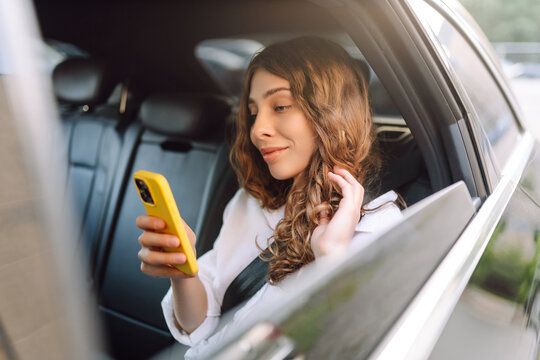 Beautiful woman with curly hair sits in car with phone in hand on sunny day. Young woman enjoys car ride and chats on smartphone. Conceptual image: transportation, blogging.