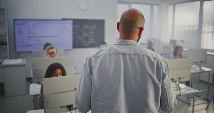 Bearded Male Teacher Leads Interactive Programming Lesson. Group of Mature Students Focus on Monitors While Listening Attentively. Concept Lifelong Learning, Innovation, and Mentorship. Back View.