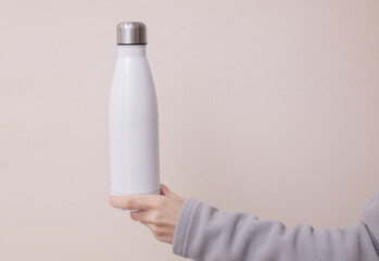 Hand in sports fleece sweatshirt holds white metal reusable bottle. Woman holds thermoson light beige background. Mockup for design. © VI-stock studio