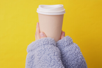 Craft eco friendly disposable cardboad cup in a female hands wearing cozy sherpa sweatshirt. Yellow background. Coffee or tea drink