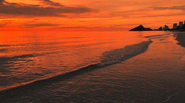 Sunset over shoreline with soft sea waves and warm red sky and Hua Hin city silhouettes on the beach