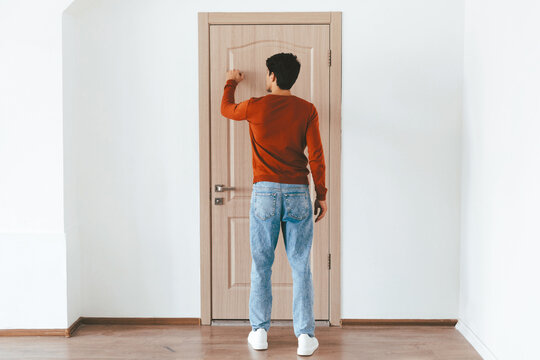 A man in a red sweater stands facing a wooden door, raising his hand to knock. The minimalist room has white walls and wooden floors, creating a calm atmosphere. - Powered by Adobe