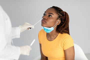 A young black woman sits calmly as a healthcare professional conducts a nasal coronavirus swab test. The setting features a neutral grey background, emphasizing the importance of health care.
