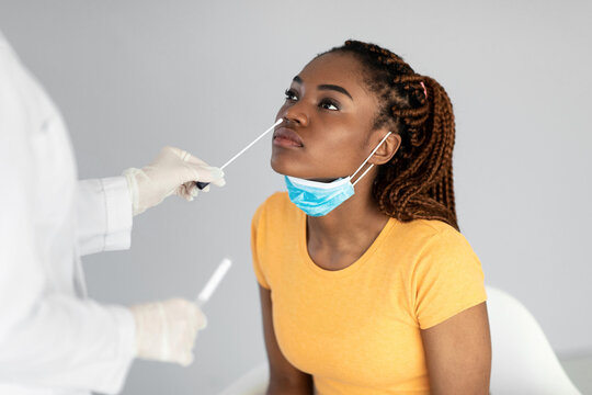 A young black woman sits calmly as a healthcare professional conducts a nasal coronavirus swab test. The setting features a neutral grey background, emphasizing the importance of health care. - Powered by Adobe