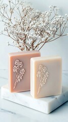 Two handcrafted soap bars, one pink and one cream, are decorated with embossed floral designs and placed on a marble surface with dried flowers.
