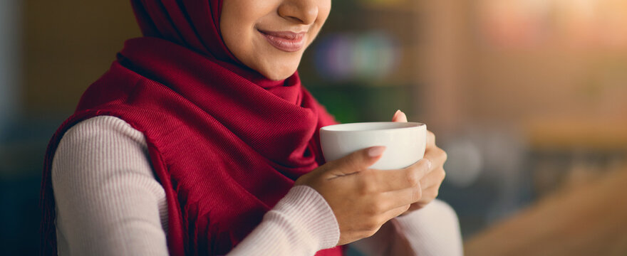 A woman dressed in a red hijab holds a white cup close to her face, smiling softly. She is indoors in a warm, inviting space, perfect for sipping on a hot drink.
