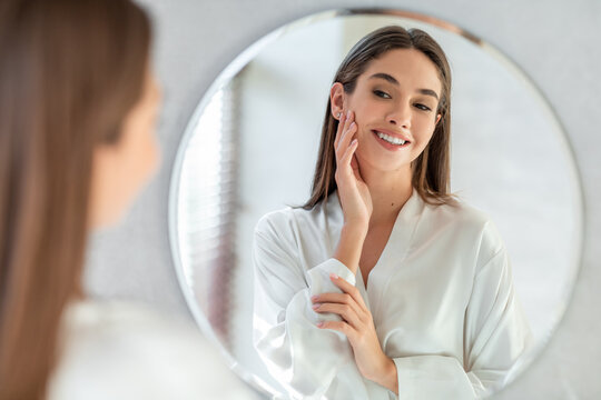 Beautiful woman in a white silk robe looks at herself in the mirror, smiling and touching her soft skin, fully engaged in her self-care routine in a well-lit space.