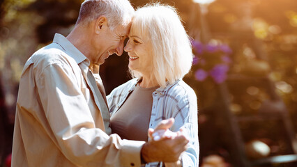 A joyful senior couple shares a dance in a vibrant garden, surrounded by blooming flowers and warm sunlight. They gaze at each other with affection, creating a heartwarming scene.