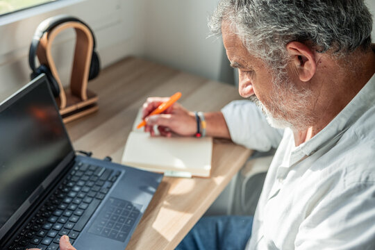 Senior man working remotely, writing notes at home office - Powered by Adobe