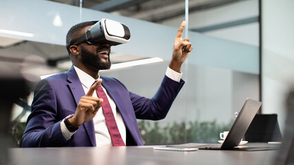 Modern technologies in business, virtual reality for enterprise concept. Black businessman sitting at workdesk in front of laptop, using VR glasses, touching copy space, office interior, panorama