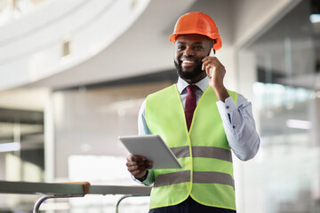 Happy african american young male architect in safety workwear and helmet having phone conversation with colleagues, using digital tablet, looking at copy space and smiling, panorama