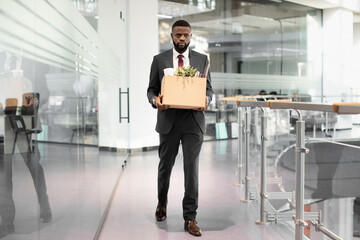 Unhappy young black man in suit employee got fired from job, walking by modern office, holding box...